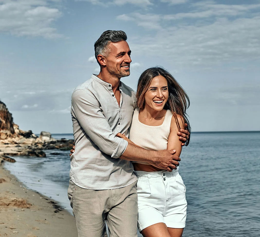 Middle aged couple holding each other lovingly while walking down the beach | Gynecomastia | NYC