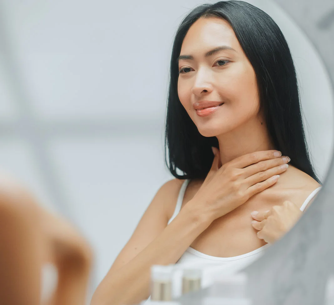 Asian woman looking in the mirror at her neck area | Neck and Chin Liposuction | NYC